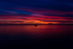 Kirkkaan punainen auringon lasku tyynellä merellä, taustalla pieni purjevene purjeet laskettuina