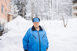 Esko Vepsäläinen poseeraa sinisessä toppatakissa ja -lakissa lumisella kerrostalon pihalla. - Sana