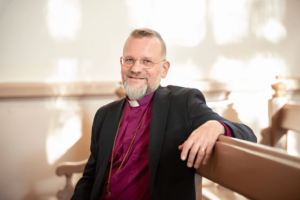 Jari Jolkkonen istuu piispan violetissa paidassa kirkon penkissä ja poseeraa kameralle. Valo tekee läikkiä valkoiseen seinään taustalla.