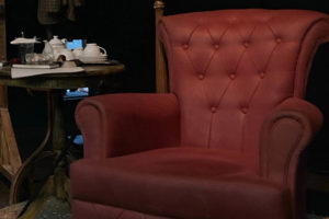 Punainen nojatuoli, jonka vierellä pieni pyöreä pöytä, jolla kahvikannu ja kupit. Hämärä valaistus.