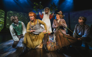 Näytelmän Anna ja Marilla istuvat kuvitteellisilla rattailla, Marilla pitää ohjia. Heidän ympärillään lapset Gilbert, Ruby, Josie ja Charlie muodostavat rattaat. Kuvassa on vauhtia.