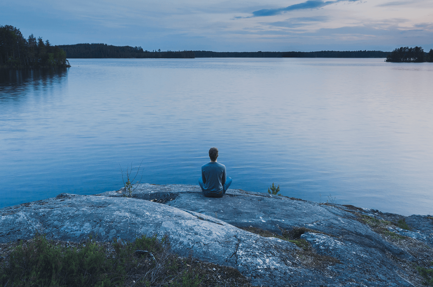 Ihminen istuu selin katsojaan kalliolla järven rannalla. Sininen, tyyni hetki. Sana