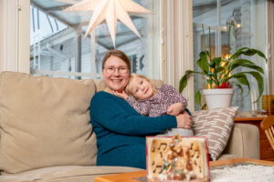 Leena Helkkula istuu sohvalla ja pitää tytärtään sylissä. Ikkunassa roikkuu tähtivalo.