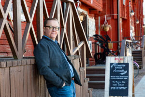 Mika Nuojua nojailee kädet taskuissa Oulun kauppatorin kahvilamakasiinin porraskaiteeseen. Taustalla punamullattu makasiinirakennus ja kahvilan liitutaulustandi.